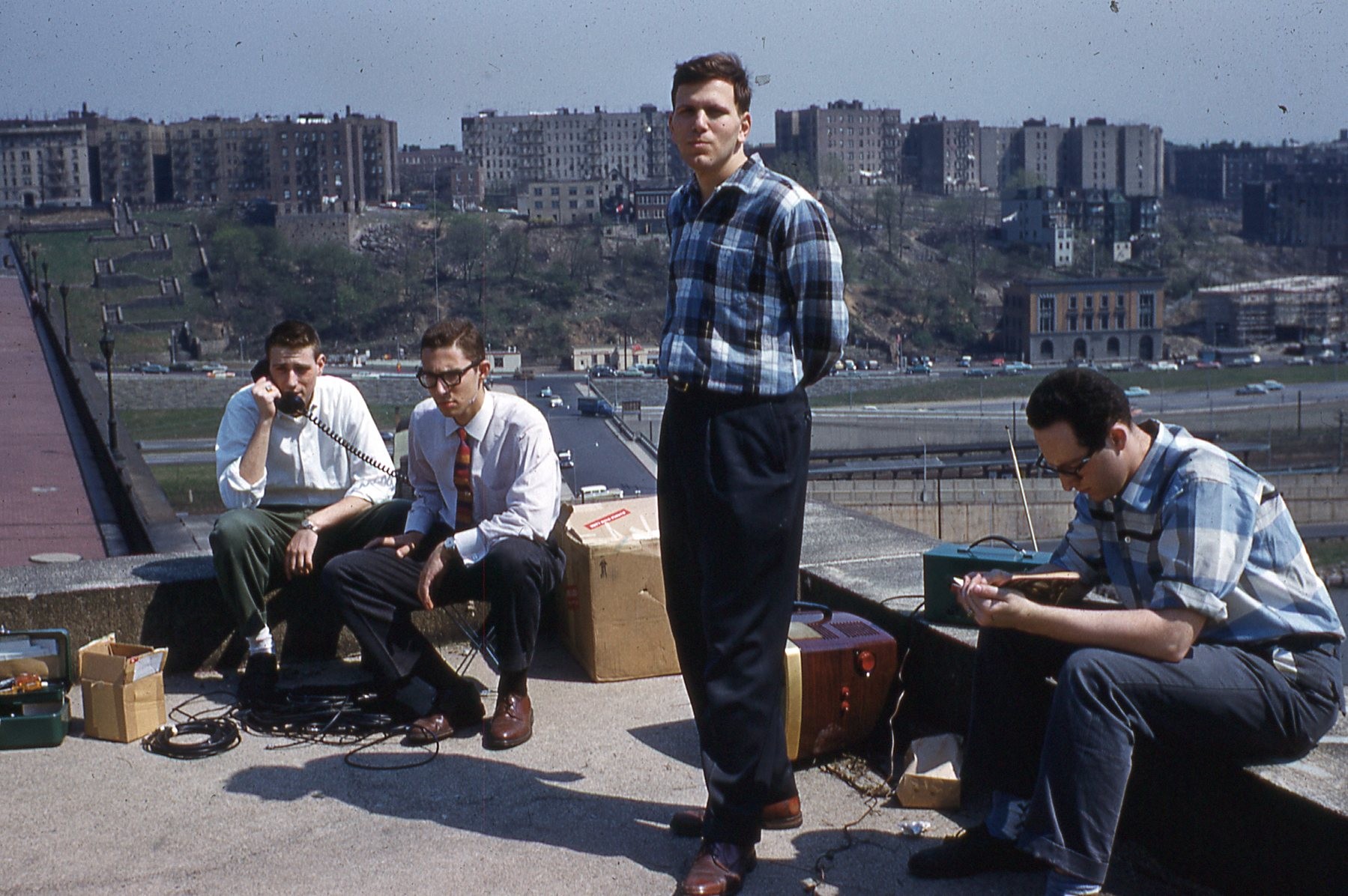 Photo of 3 men seated and one standing on rooftop with distant buildings in background