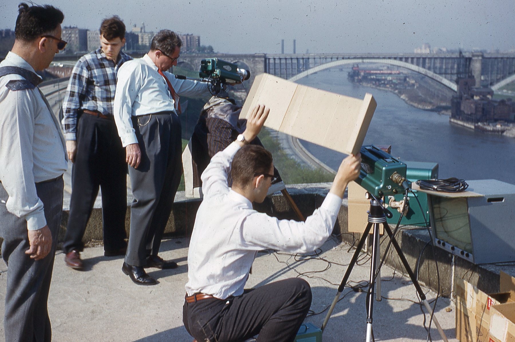photo of 4 men standing on rooftop looking at TV cameras with river and bridge in distant background